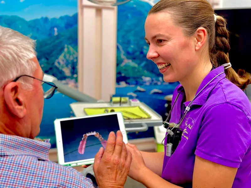 Zahnärztin zeigt einem älteren Patienten ein 3D-Modell des Gebisses auf einem Tablet und bespricht die Behandlung