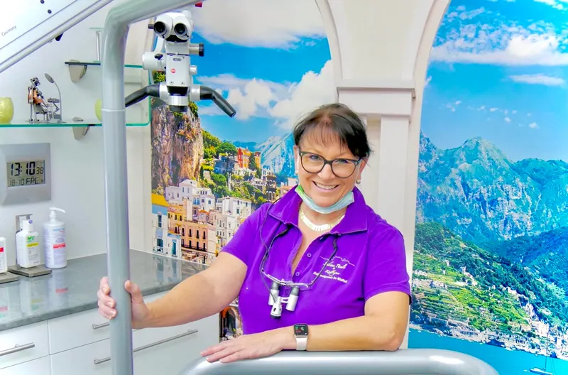 Zahnärztin im Behandlungszimmer mit OP-Mikroskop und Wandbild einer Küstenlandschaft